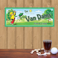 Personalized golf sign featuring a cartoon golfer in a yellow hat and red pants, swinging a golf club and sending a golf ball flying into a blue sky. The scene includes a clump of green grass and a tee, with customizable text displaying your family name. Perfect for adding a humorous touch to any golfer's space
