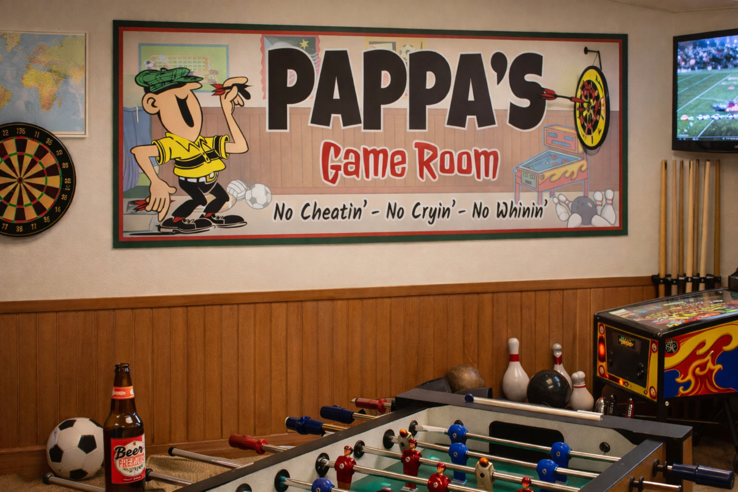 Pappa’s Game Room sign mounted on a game room wall above a foosball table with dartboard, TV, and arcade machines