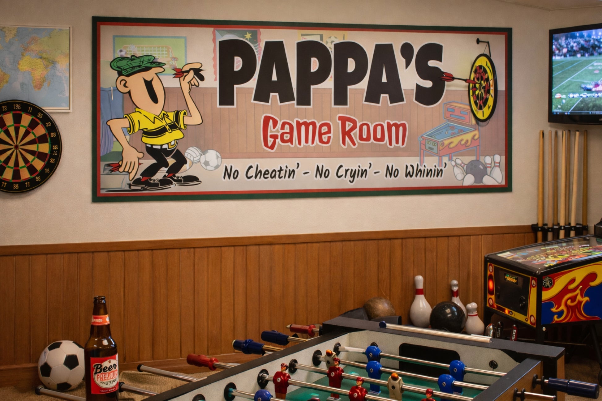 Pappa’s Game Room sign mounted on a game room wall above a foosball table with dartboard, TV, and arcade machines