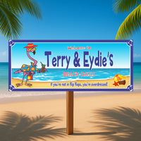 Personalized beach oasis sign with flamingo holding a drink, displayed on the sand with ocean and palm trees in the background.
