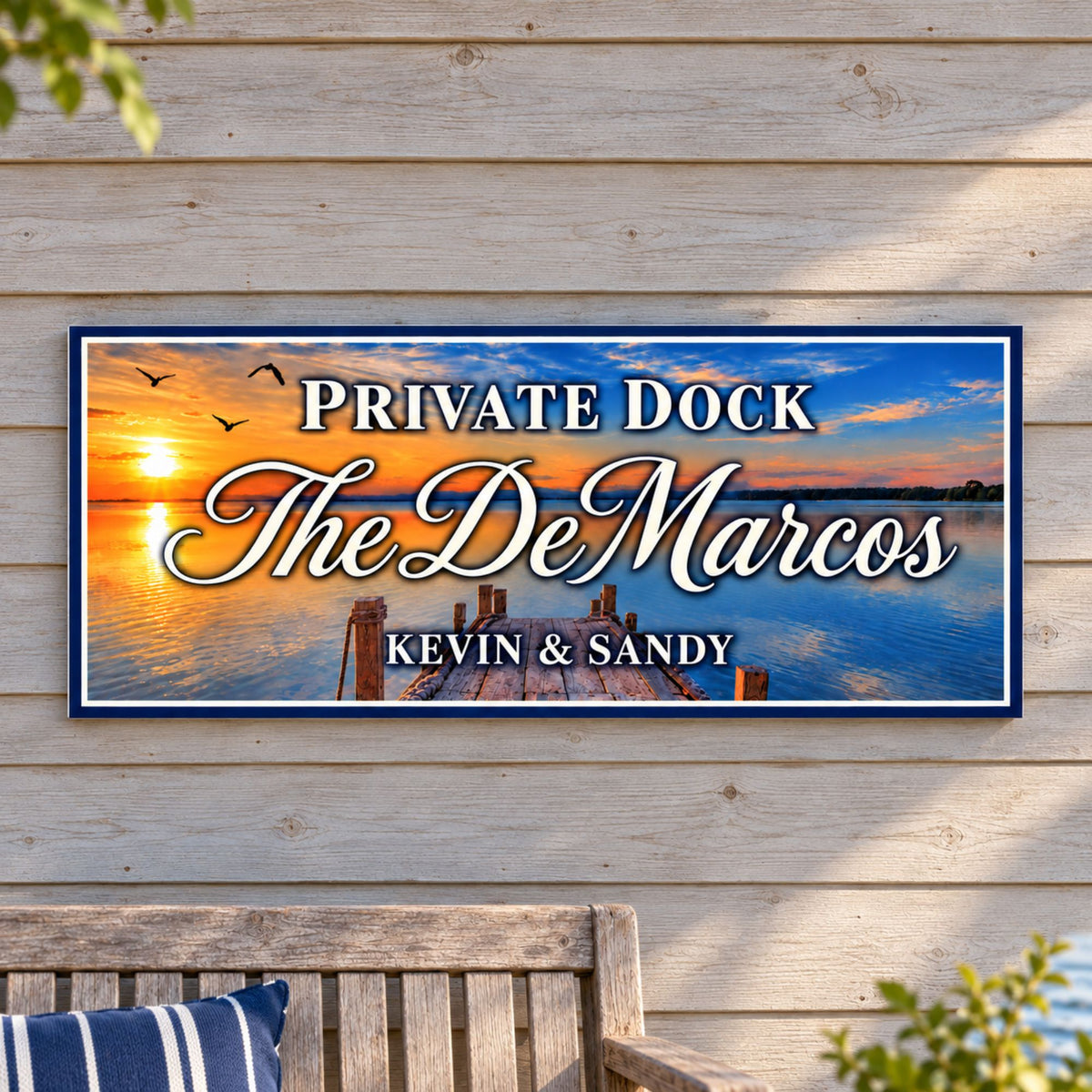 Custom sunset dock sign mounted on pale wood siding above an outdoor bench with a navy striped cushion.