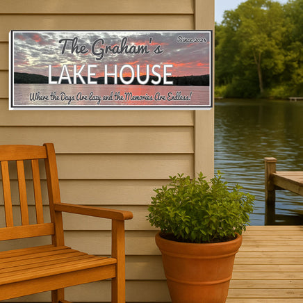 Personalized lake house wall sign mounted on an exterior siding near a dock, wooden bench, and potted plant with calm lake and trees in the background, customizable family name and phrase