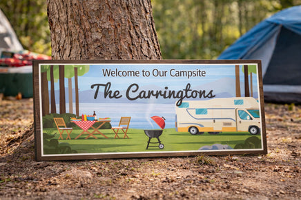 Personalized camping sign reading “Welcome to Our Campsite – The Carringtons,” shown in a 28x11 rectangular shape leaning against a tree at a campground, with an RV, picnic table, and grill in the background.