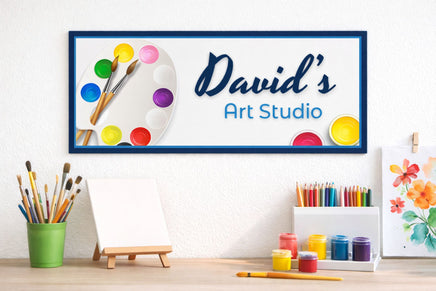 Personalized art studio sign on wall above an art table with paintbrushes, pencils, and paint jars, showing a custom name design with colorful paint palette