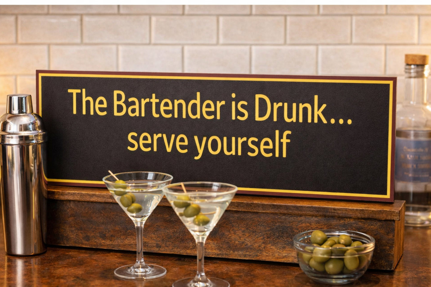 Bartender humor sign resting on a bar top with martini glasses, cocktail shaker, and olives in a home bar setting