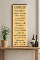 Vertical “Motto to Live By” wall sign displayed on a wooden shelf with a plant and wine bottles, humorous quote about living life to the fullest.