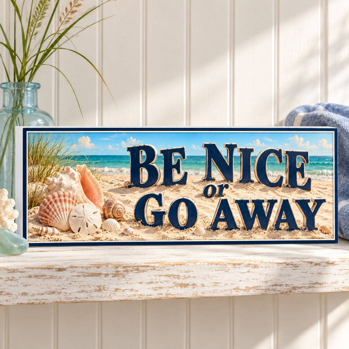 Be Nice or Go Away sign displayed on a whitewashed coastal shelf beside a sea-glass vase and blue linen.