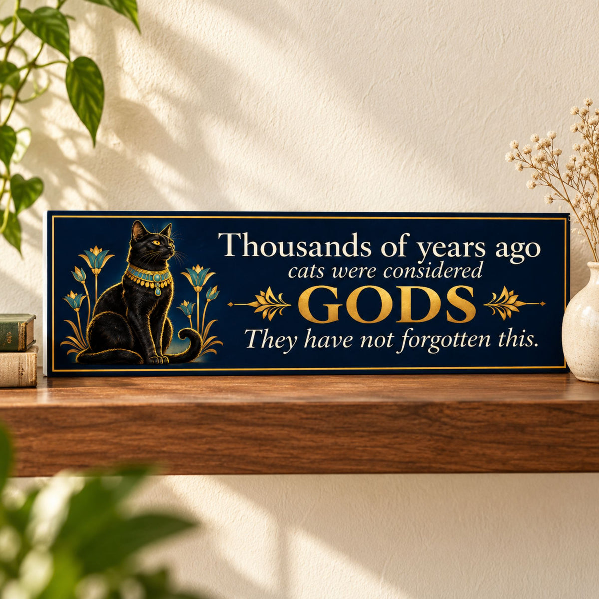 Navy and gold cat quote sign displayed on a walnut shelf, featuring a regal black Egyptian cat and the words “Thousands of years ago cats were considered gods. They have not forgotten this.”