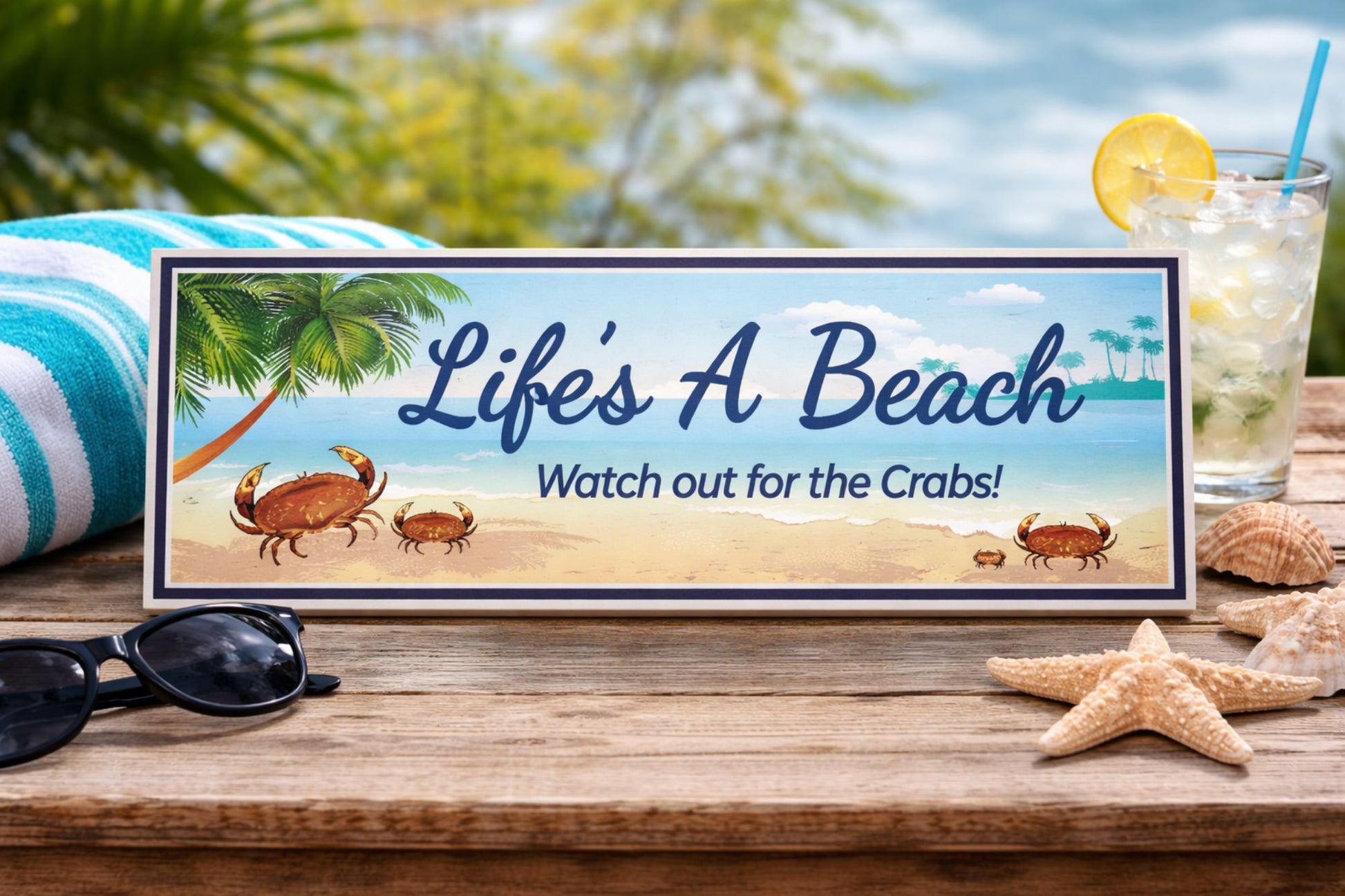 Life’s A Beach beach sign displayed on wooden table with towel drink and seashells coastal shelf decor scene