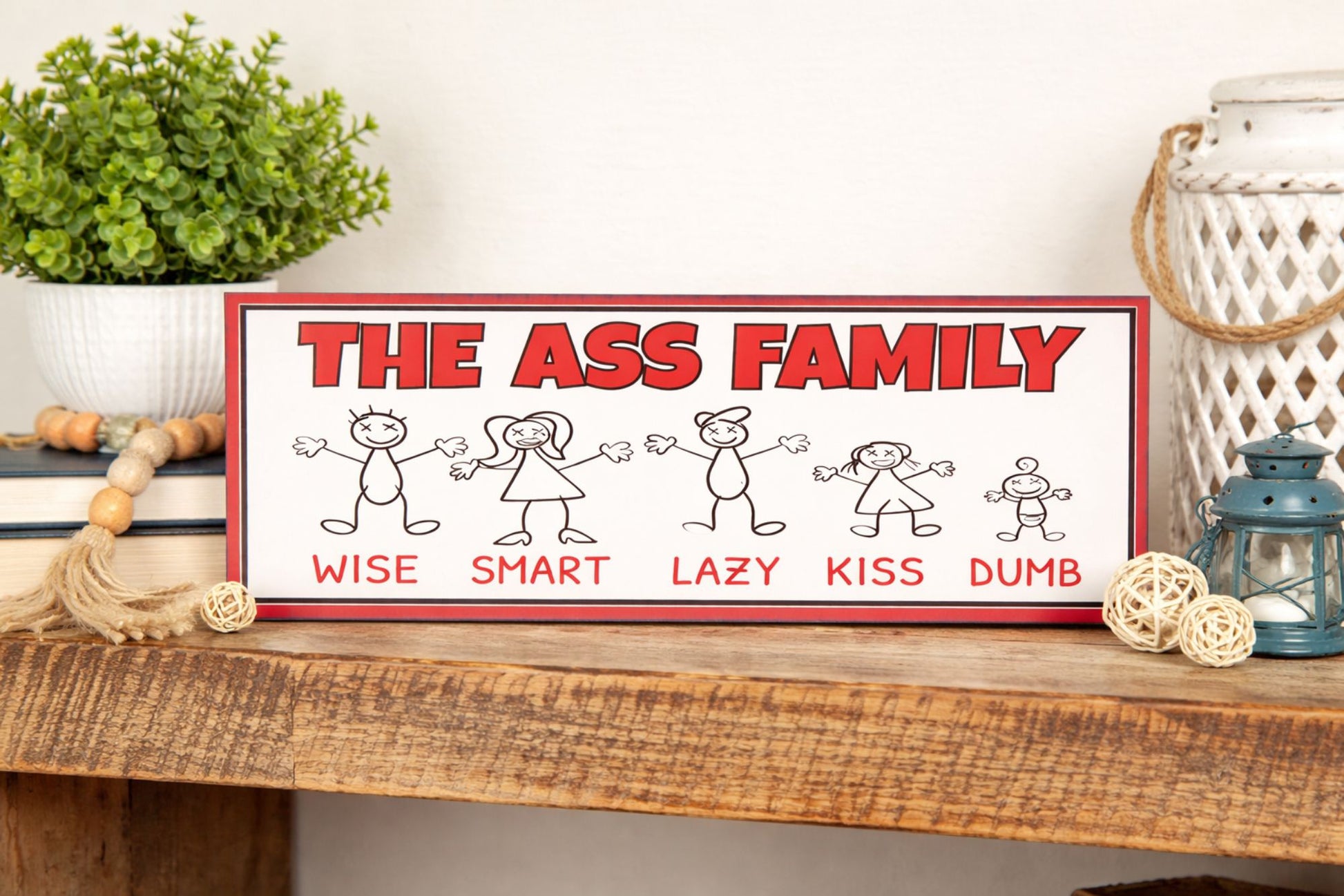 funny ass family sign displayed on rustic wood shelf with plant books lantern and decor close up horizontal novelty sign