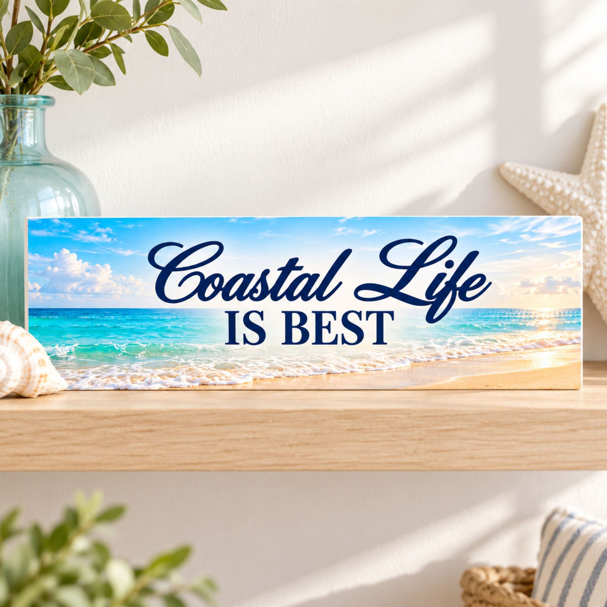 Coastal Life Is Best beach sign displayed on a wooden shelf beside a blue glass vase and seashells, with turquoise ocean waves, sandy shore, and navy lettering.