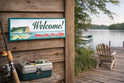Rustic lakeside cabin scene with a fishing welcome sign mounted on wood siding, showing a bass illustration and the phrase “Drop a Line – Stay a While,” next to a dock, fishing gear, and Adirondack chair overlooking the lake.