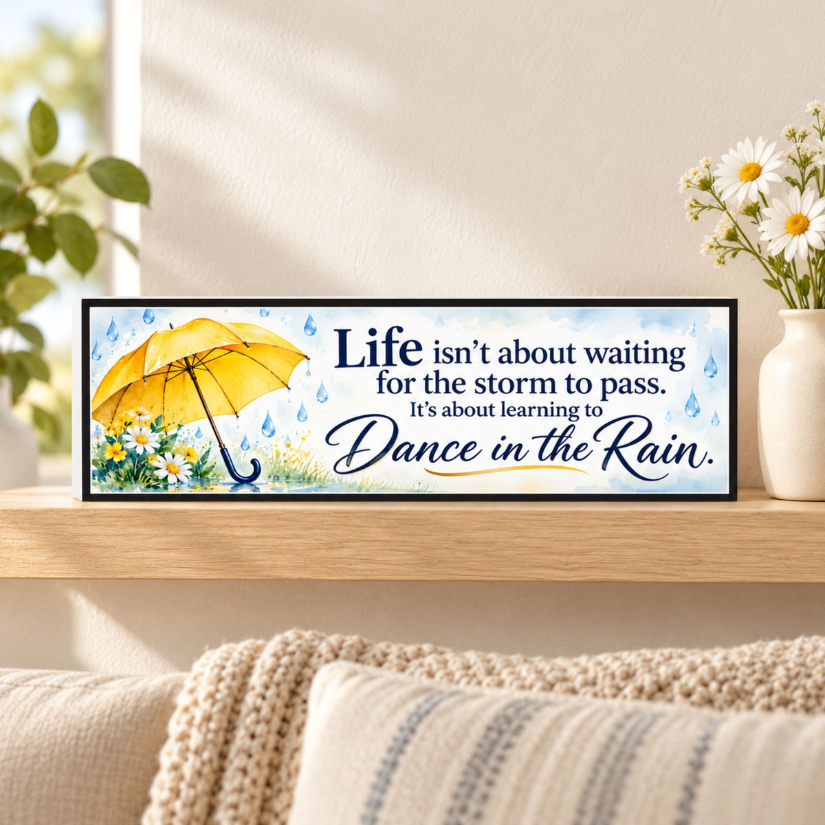 Dance in the Rain inspirational sign with a yellow umbrella, daisies, and black border, displayed on a wooden shelf beside white flowers.