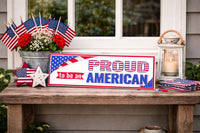 Patriotic “Proud to be an American” sign displayed on a rustic front-porch table with small American flags, flowers, and a lantern.