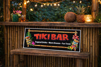 Tiki Bar novelty sign with bamboo border and colorful tiki masks leaning against a bamboo bar, surrounded by string lights, tropical drinks, a coconut, and a pineapple for a warm backyard tiki atmosphere.