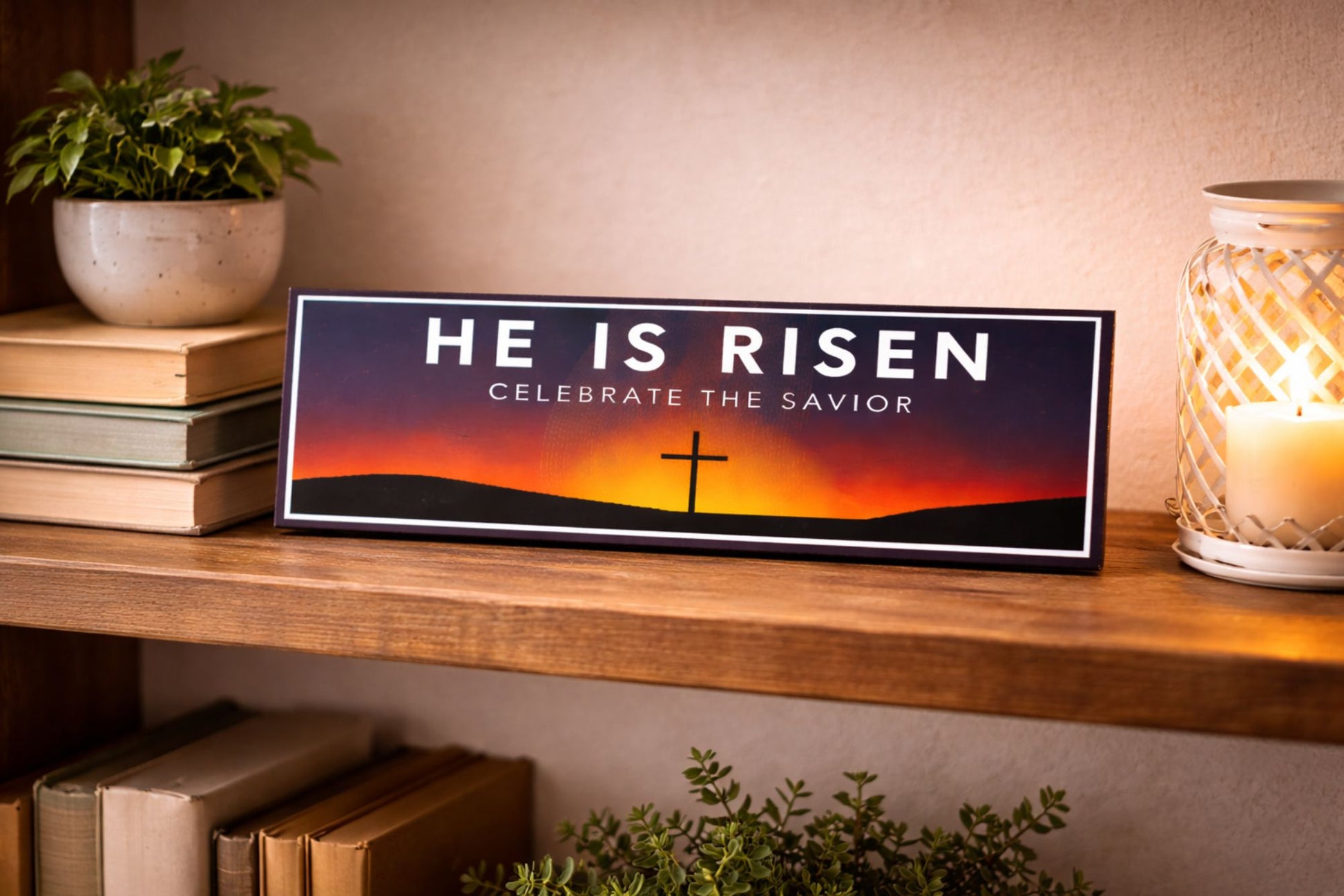 he is risen sign displayed on shelf with candle books and plant in home setting