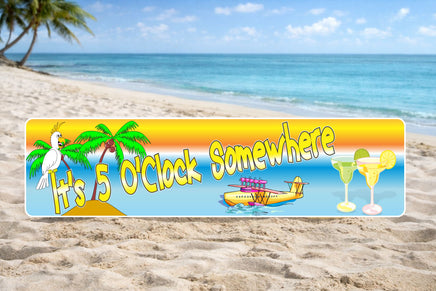 It’s 5 O’Clock Somewhere tropical sign displayed on a sandy beach with ocean and palm trees in the background