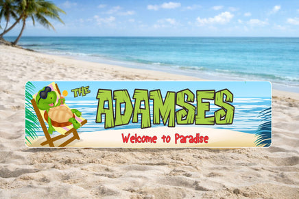 Personalized aluminum beach sign sitting in the sand with ocean waves behind it, featuring a turtle in sunglasses and the phrase Welcome to Paradise.