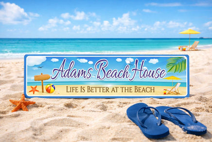 Personalized aluminum beach house sign placed in the sand by the ocean, with flip-flops, starfish, and beach umbrella in the background.